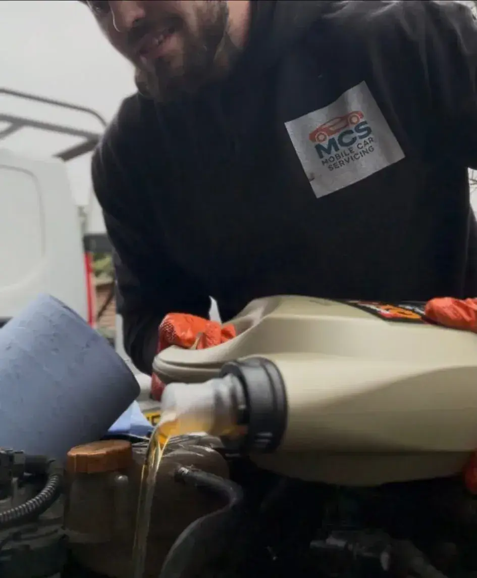 Mechanic in MCS Mobile Car Servicing branded hoodie pouring fresh oil into an engine